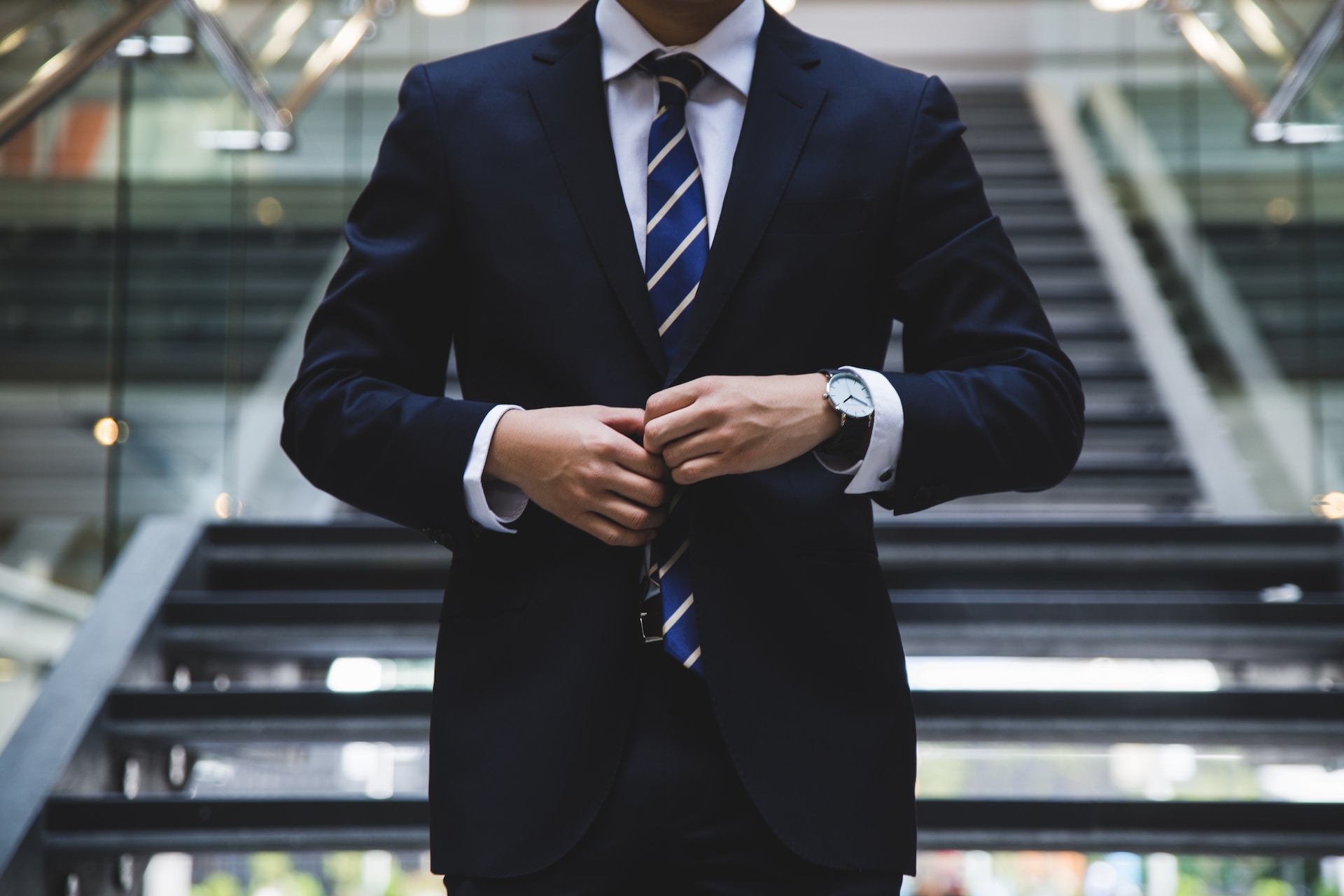 Caucasian man in a dark blue suit buttoning it up with a blue and white stripe tie wearing a watch. . Behind him is a blurred set of stairs in what appears to be a large office building.