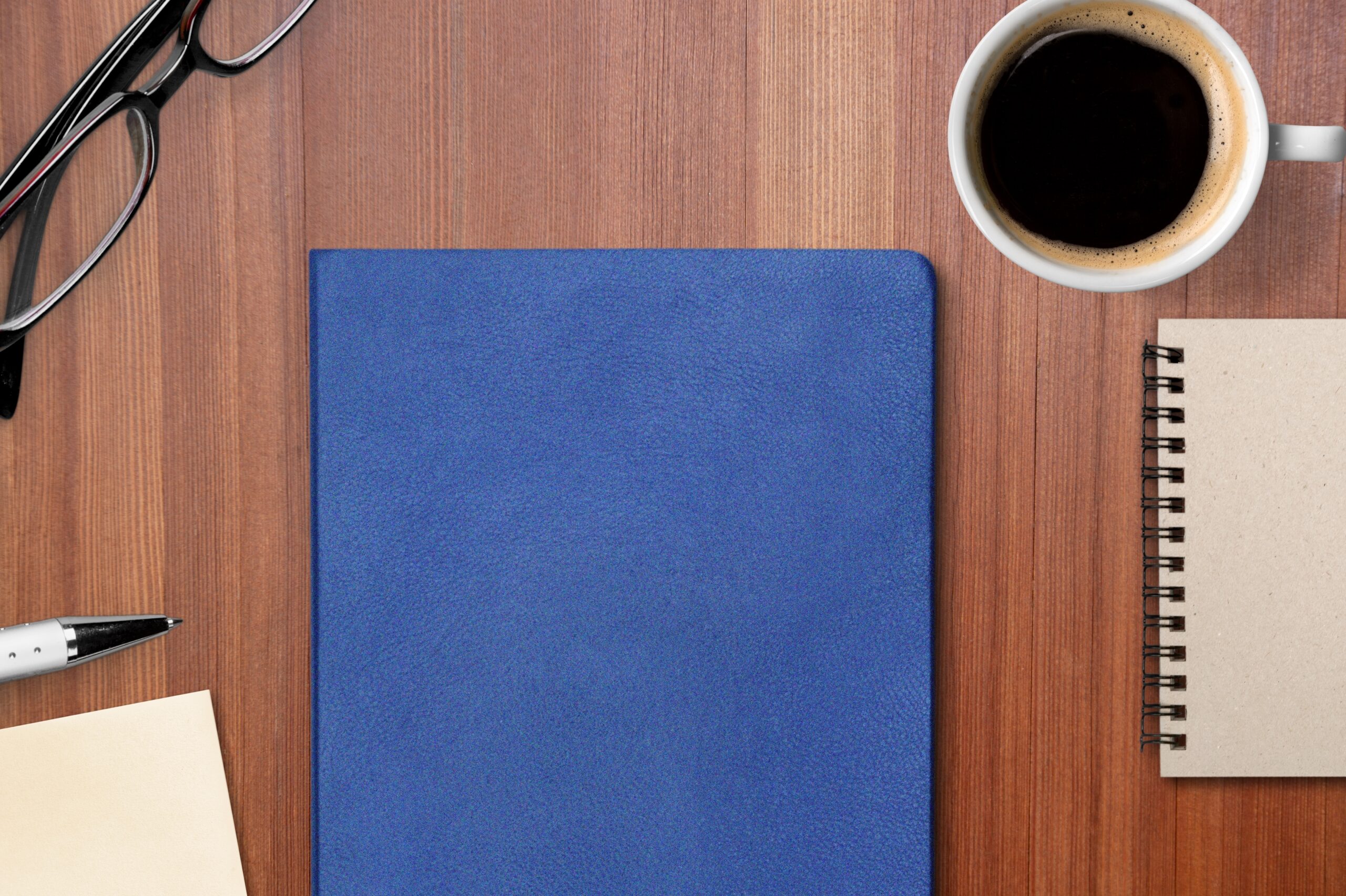 Blue book on a light wooden desk with a cup of coffee a smaller notebook with black ring binding and a peace of paper, a pen, and eye glass with black rim. 